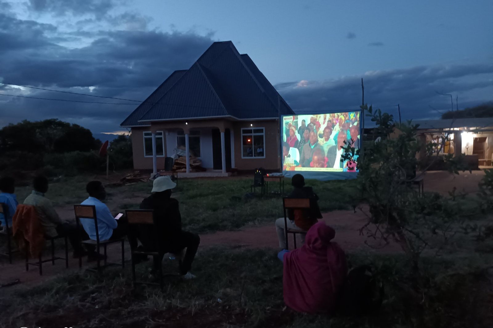 Villagers seated outdoors at dusk watching a programme projected onto a screen